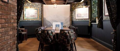 A dining area with a wooden table and floral upholstered chairs, brick walls, and framed pictures on the walls.