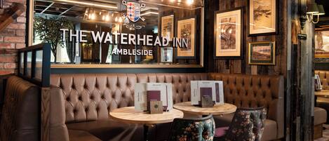 A cozy interior with a sign reading "The Waterhead Inn Ambleside," featuring a tufted leather sofa, floral chairs, and framed artwork on the walls.
