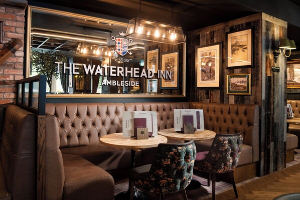 A cozy interior with a sign reading "The Waterhead Inn Ambleside," featuring a tufted leather sofa, floral chairs, and framed artwork on the walls.