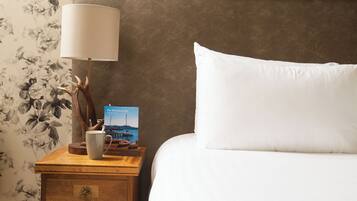 Ein ordentlich bezogenes Bett mit einem weißen Kissen und einem Nachttisch daneben. Auf dem Nachttisch stehen eine Lampe mit weißem Schirm, eine Tasse und ein Buch mit dem Titel "The Waterford Inn".