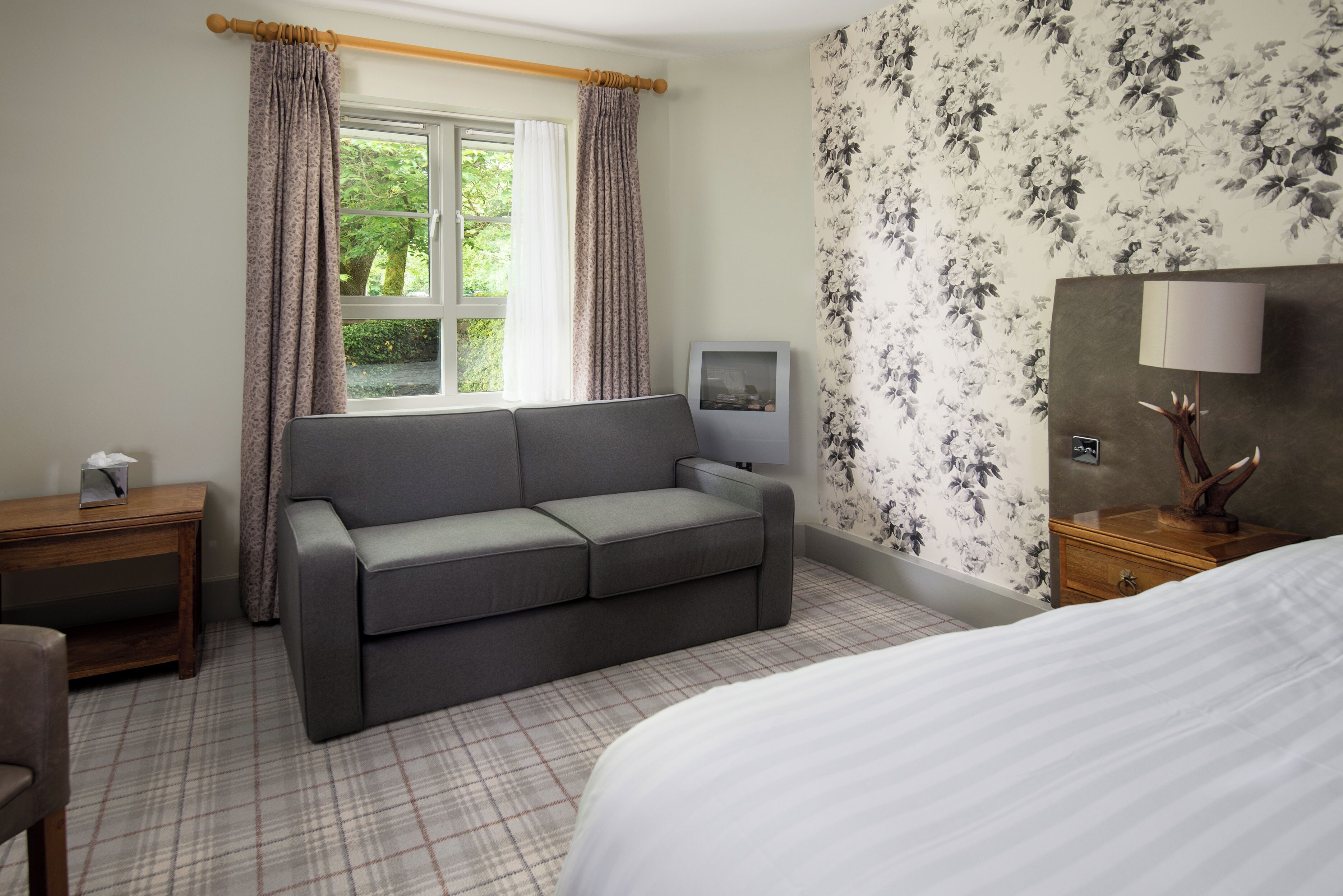 Habitación de hotel con un sofá gris, una mesita de noche de madera con una lámpara, una cama con ropa blanca y una ventana con cortinas, además de un papel tapiz con estampado floral.