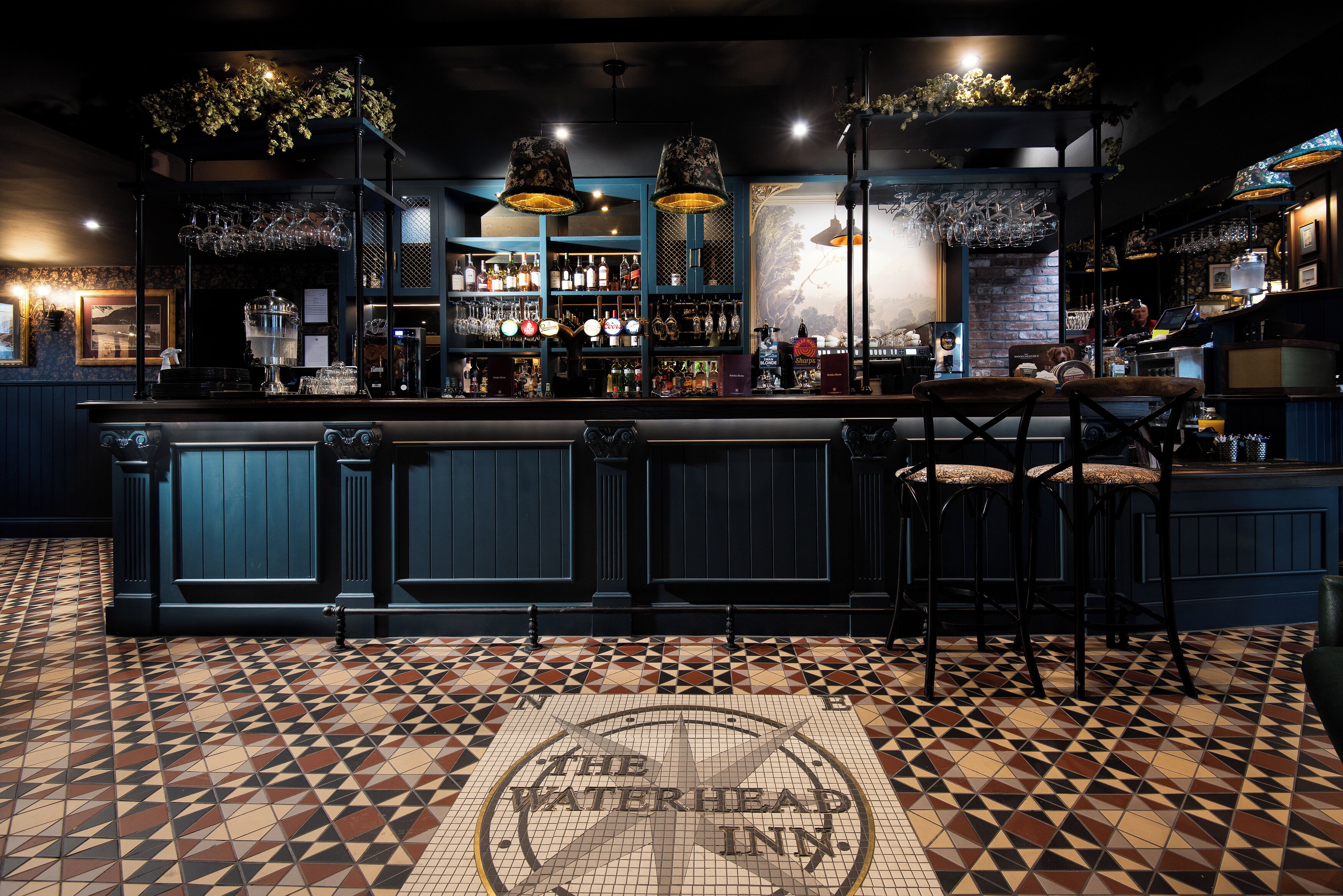 Zona de bar con suelo de baldosas, taburetes y mostrador con una variedad de botellas y vasos.