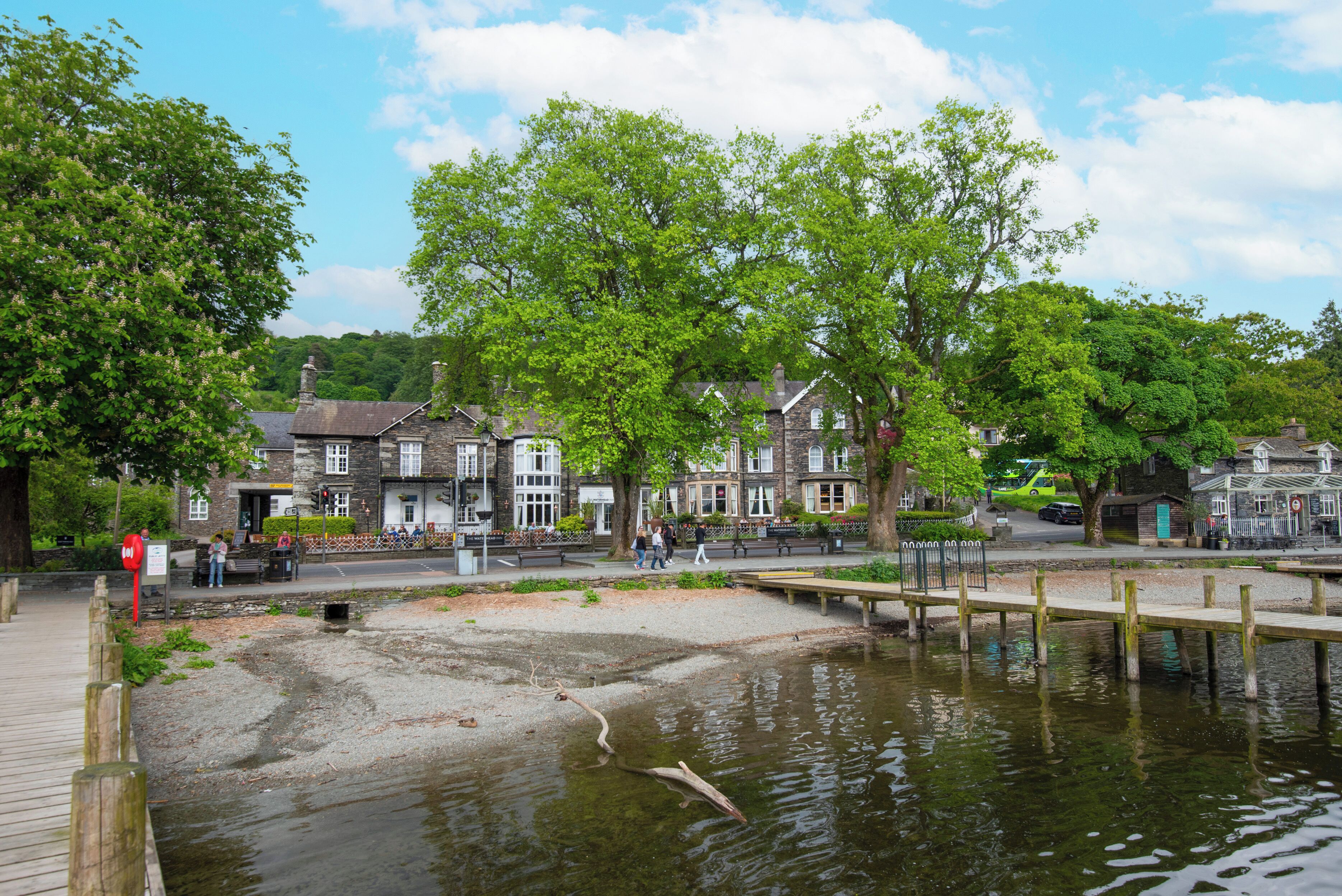 Un paseo junto al río con un muelle de madera, edificios de piedra y una zona ajardinada.