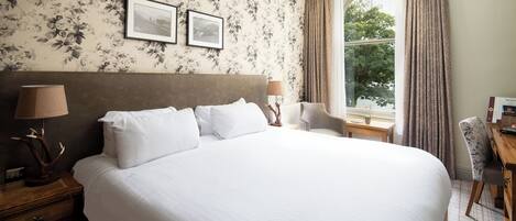 A neatly made bed with white linens, a wooden nightstand with a lamp, a chair, and a desk with a lamp. Two framed pictures are on the wall above the bed.