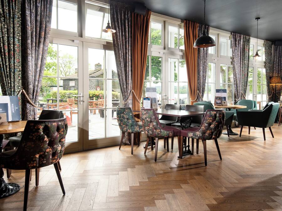 Un comedor amplio con suelo de madera, sillas tapizadas con motivos florales y ventanas grandes con cortinas.