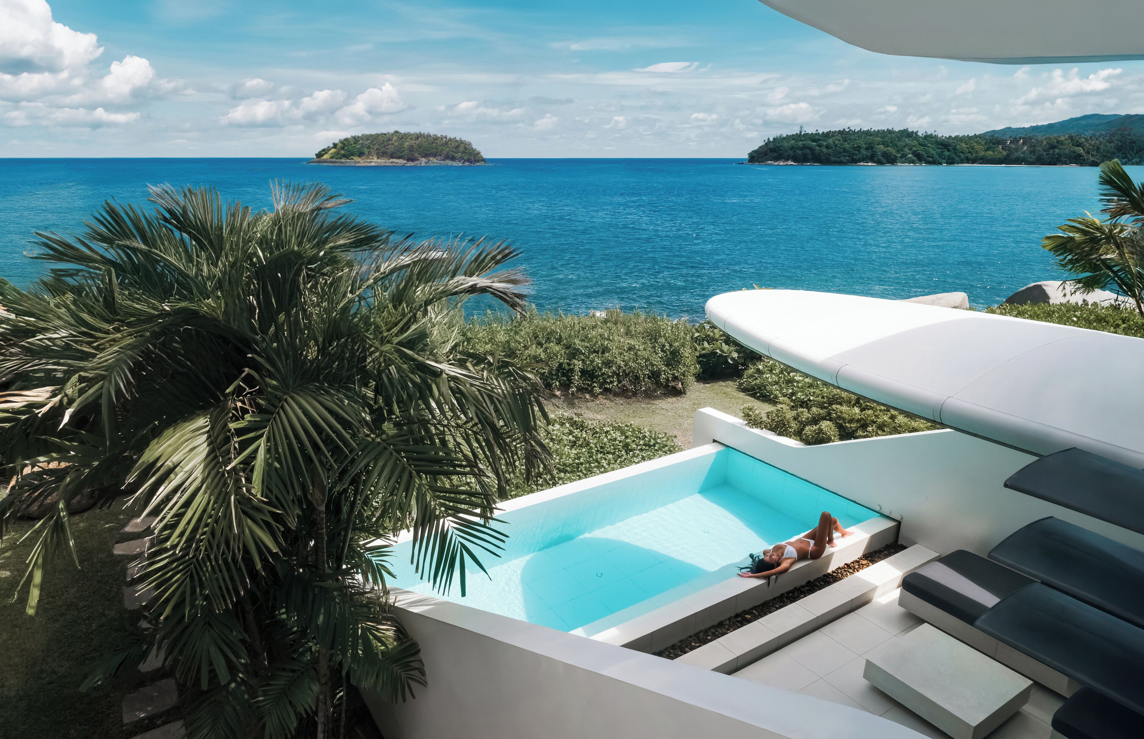 A pool with a view of the sea, surrounded by palm trees and a white structure.