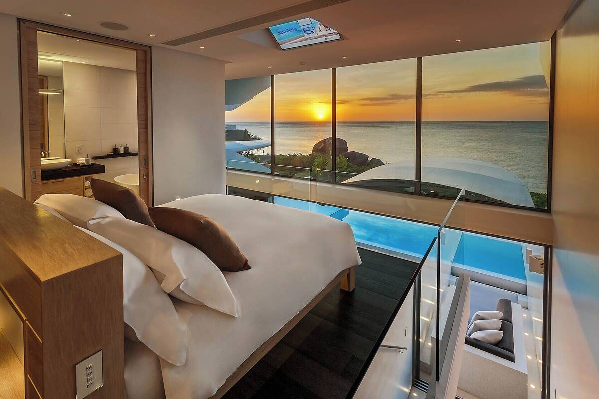 A bedroom with a large bed, a bathroom with a sink, and a view of the ocean at sunset.