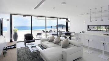 A modern living room with a large window overlooking a beach, a white sofa, a dining area, and a kitchen with pendant lights.