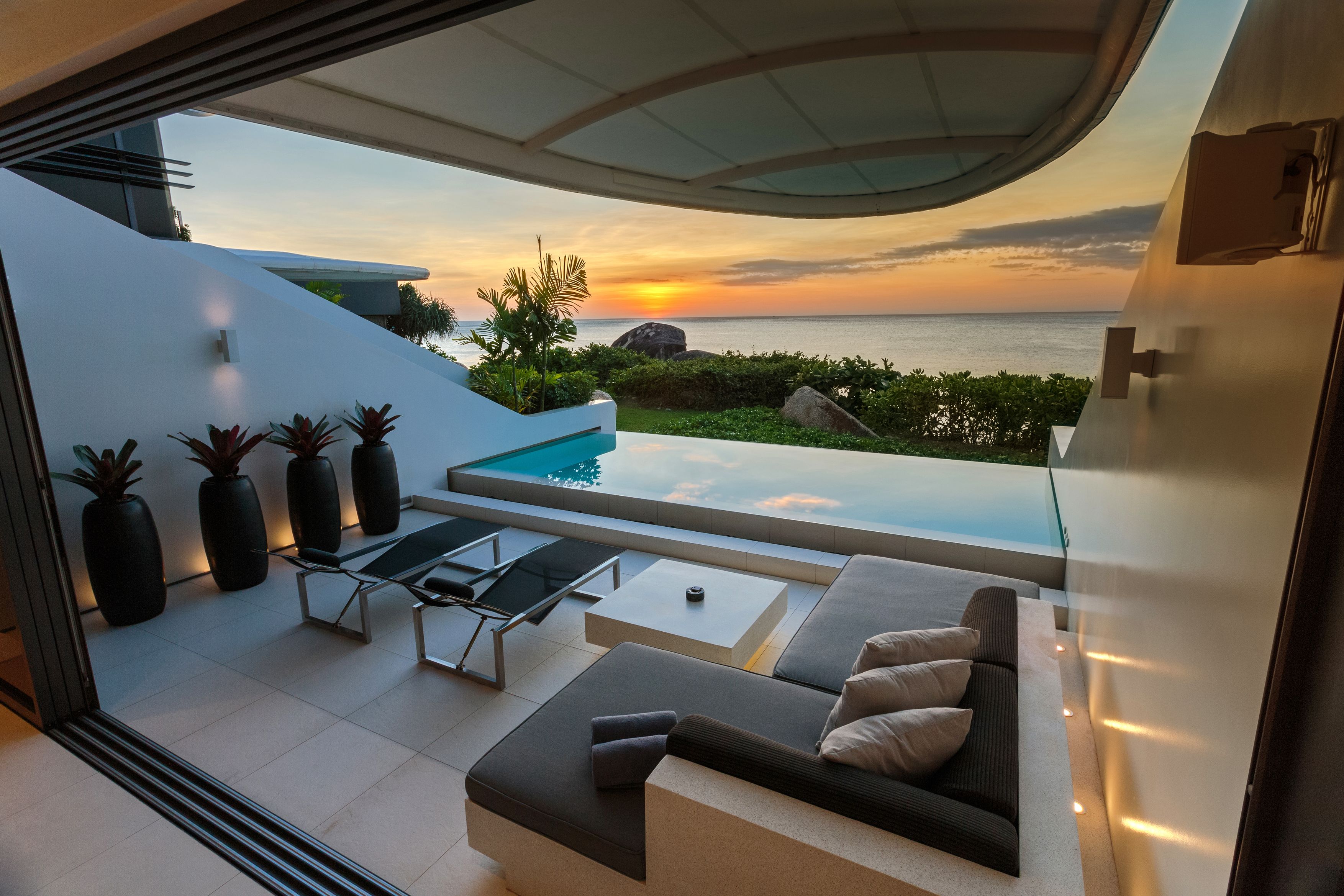 A modern outdoor living area with a pool, seating, and a view of the ocean at sunset.