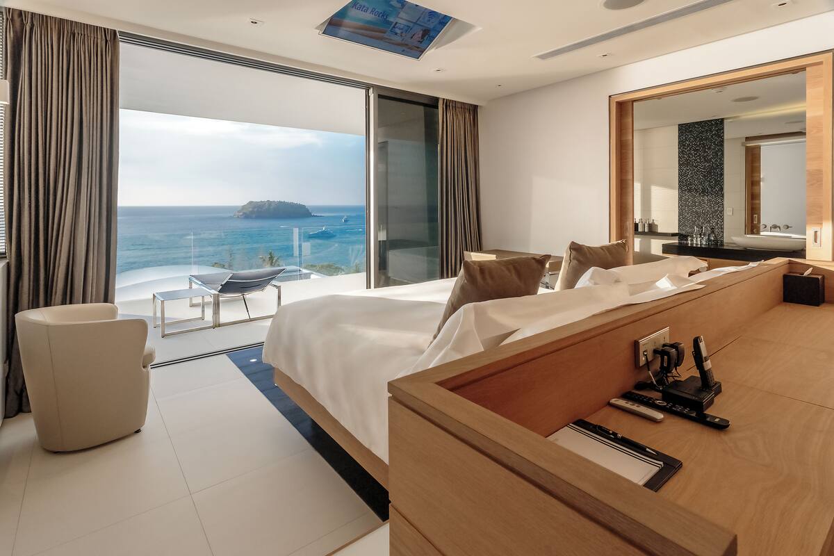 A modern hotel room with a large bed, a wooden desk, a chair, a balcony with a view of the sea, and a large mirror.