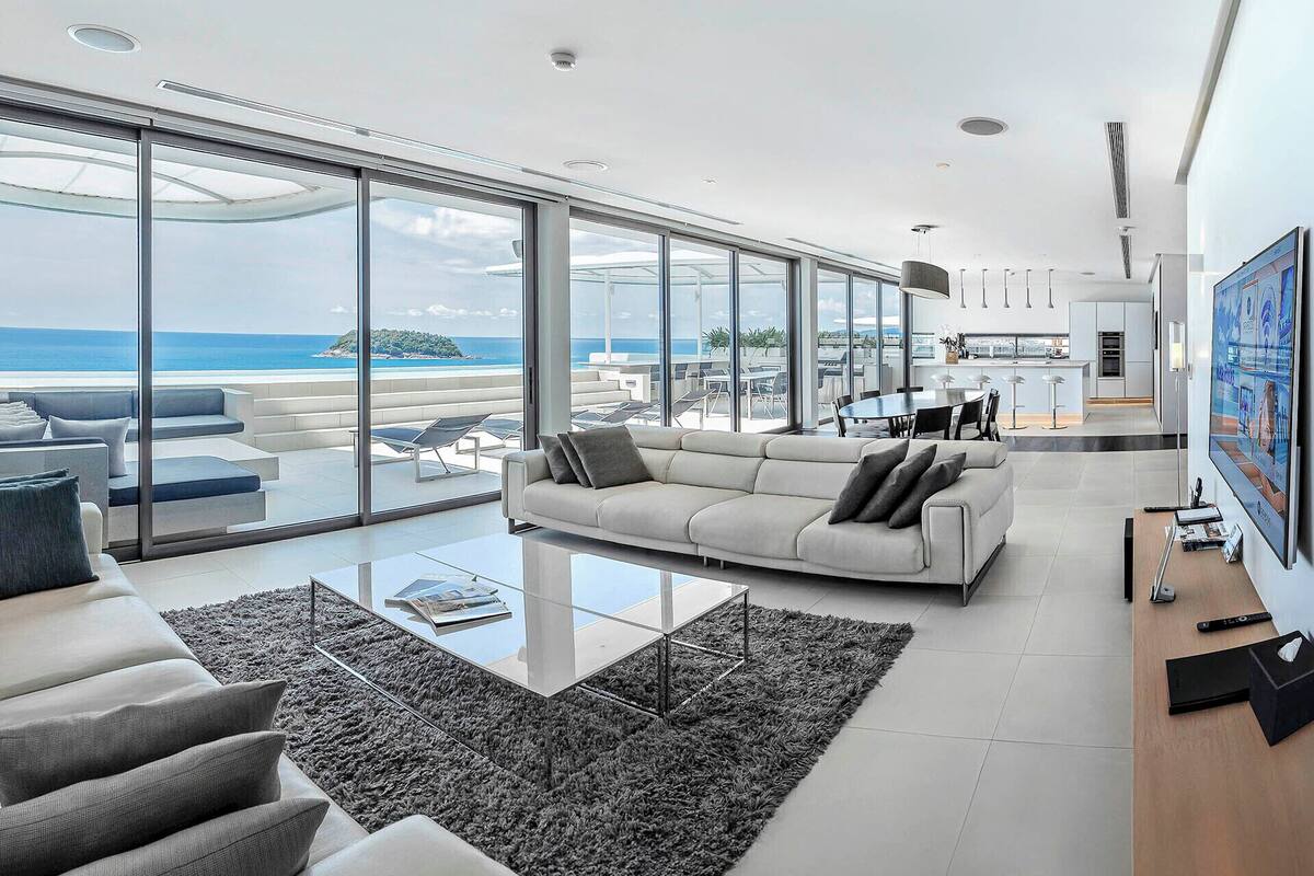 A modern living room with a large glass wall offering a view of the ocean and a balcony with outdoor seating.