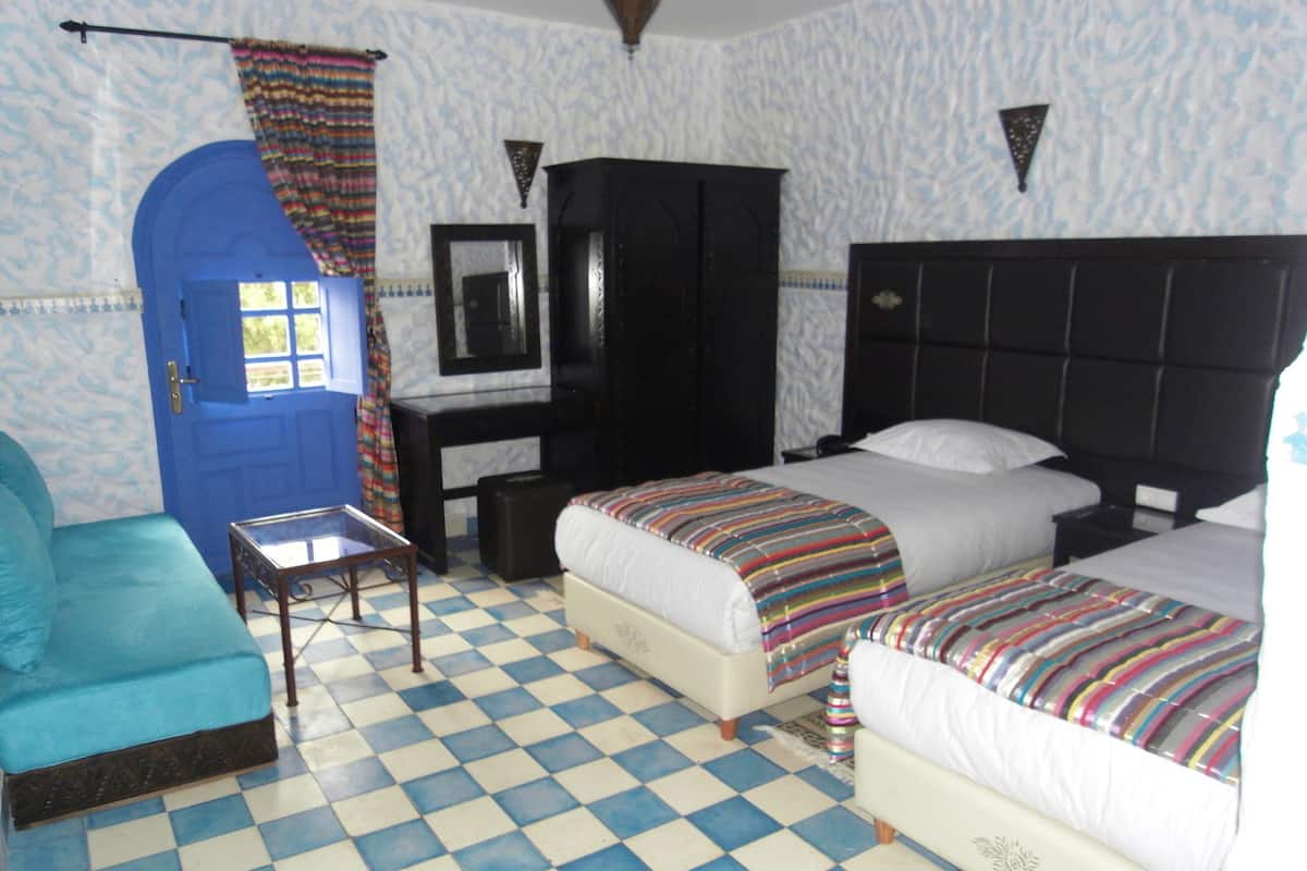 A room with a checkered floor, a blue door, a bed with a striped blanket, a bedside table, a mirror, and a wardrobe.
