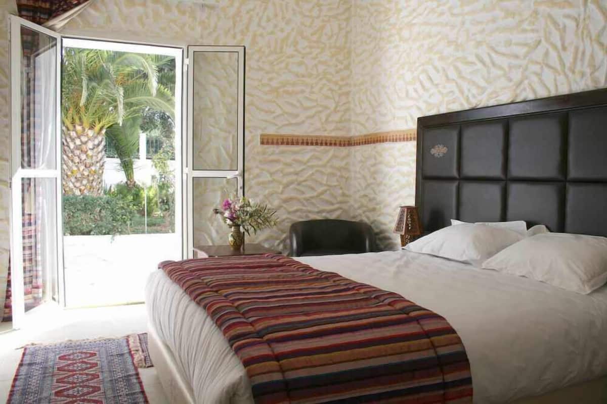 A bedroom with a bed, a chair, a desk, and a view of palm trees through a glass door.