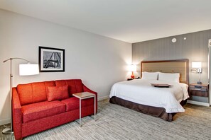 A hotel room with a red sofa, a bed with white bedding, a small side table, and a framed picture on the wall. - Hampton Inn & Suites Detroit/Troy (Troy)