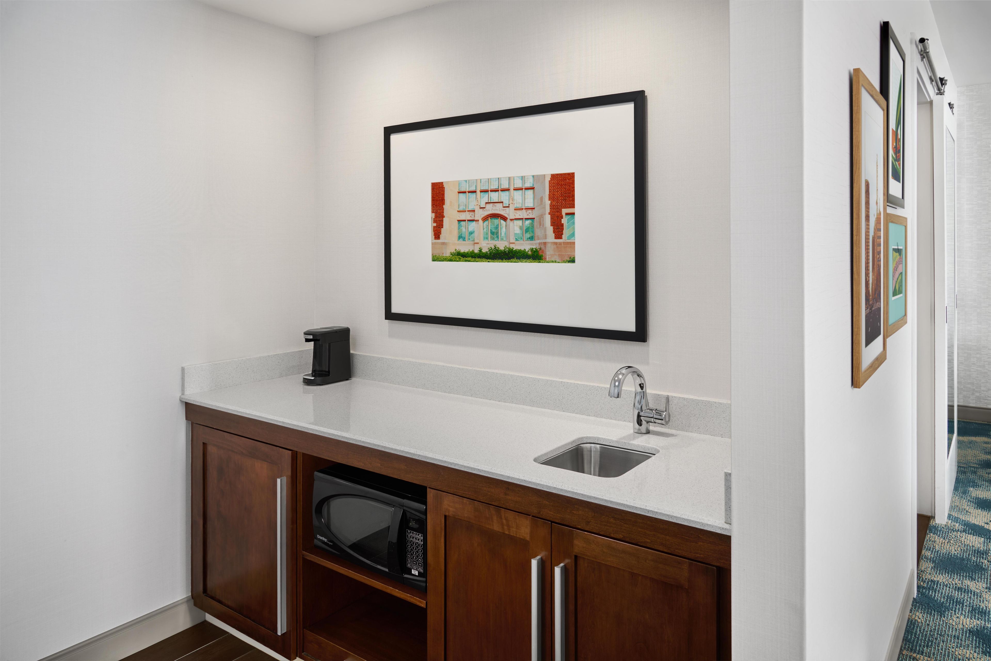 A kitchenette with a microwave, sink, and cabinet, a framed picture on the wall, and a patterned carpet.