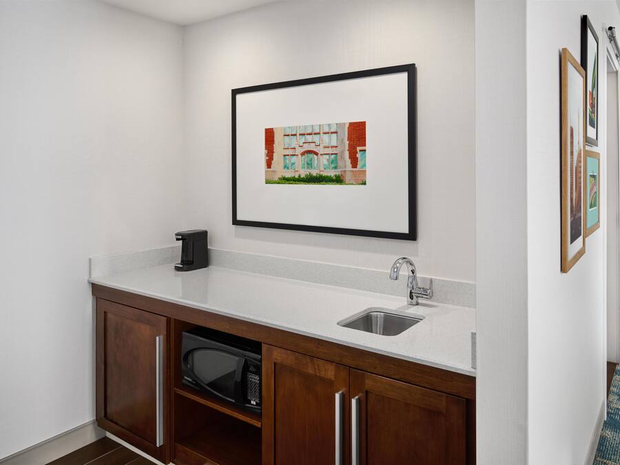 A kitchenette with a microwave, sink, and cabinet, a framed picture on the wall, and a patterned carpet.