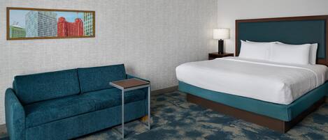 A hotel room with a bed, a sofa, a small table, a lamp, and a cityscape painting on the wall.
