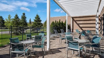 Une terrasse extérieure avec des chaises et des tables, un espace abrité et, au fond, un bâtiment en briques.
