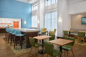 Un hall d’hôtel moderne avec un buffet pour le petit-déjeuner, des chaises vertes et jaunes, des tables en bois et de grandes fenêtres.