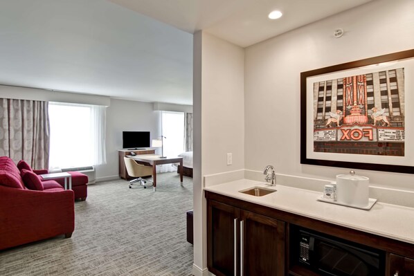A hotel room with a red sofa, a desk with a chair, a TV, a large framed picture, and a kitchenette with a sink and microwave. - Hampton Inn & Suites Detroit/Troy (Troy)
