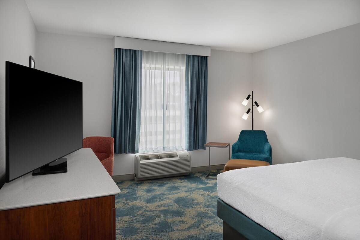 A hotel room with a bed, a TV, a desk, a chair, a lamp, and a window with curtains.