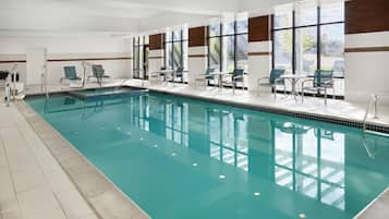 An indoor swimming pool with clear water, surrounded by lounge chairs and tables, large windows, and a tiled floor.