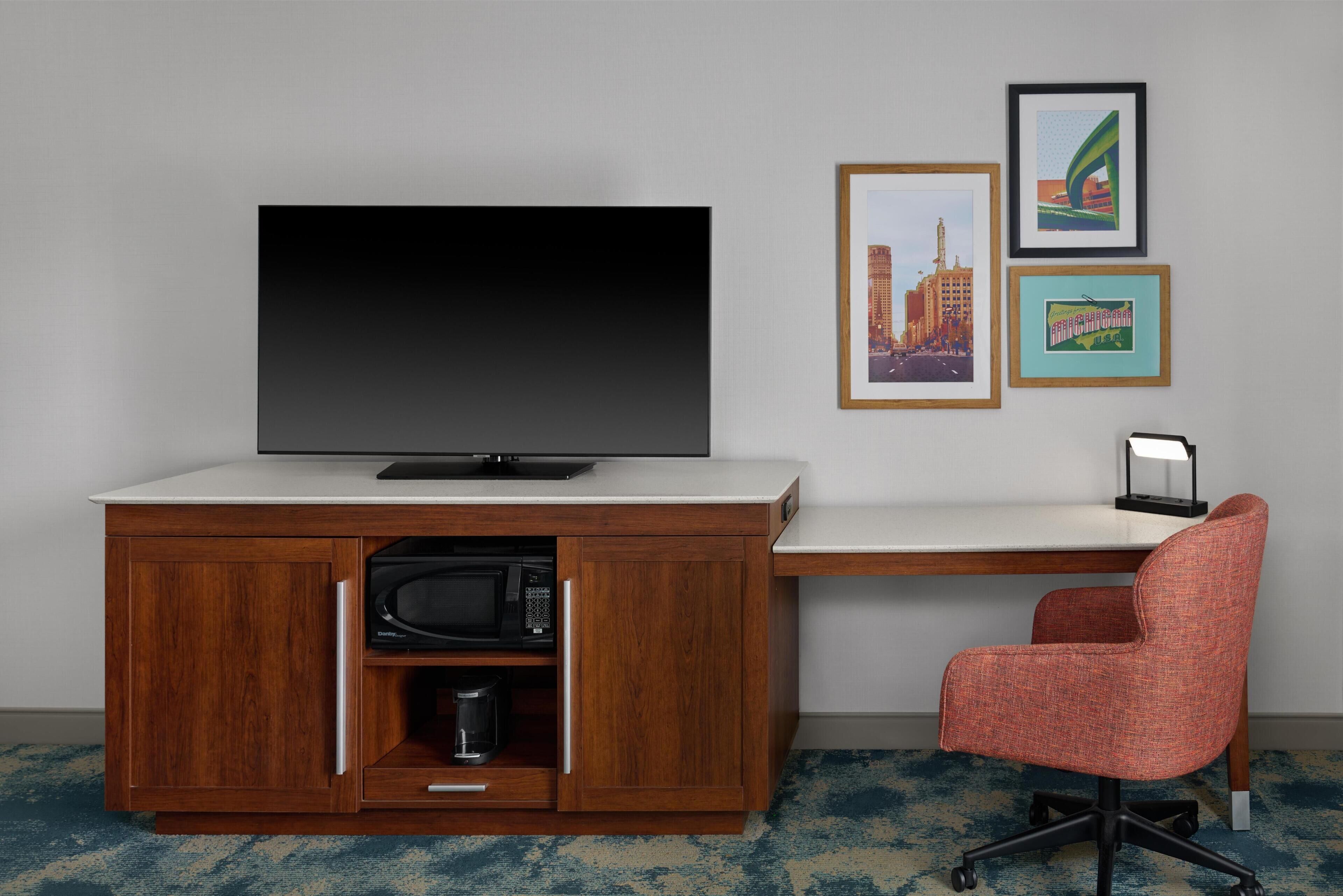 Una habitación con televisión sobre un mueble de madera, un escritorio, una silla y cuadros en la pared.
