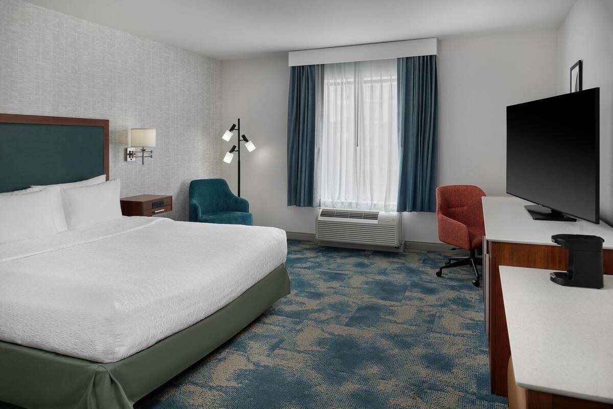 A hotel room with a large bed, a desk with a TV, a chair, a lamp, and an air conditioning unit.
