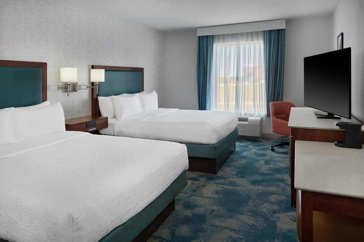 A hotel room with two beds, a desk, a chair, a television, and a large window with curtains.