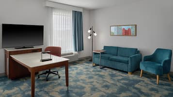A hotel room with a flat-screen TV, a desk, a blue sofa, a blue armchair, a floor lamp, and a cityscape painting.