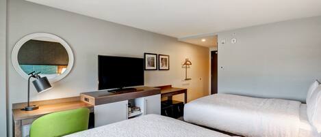 A hotel room with two beds, a desk with a computer, a TV, and a round mirror.