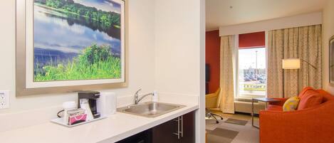 A hotel room with a kitchenette, a large window with curtains, a sofa, and a painting of a landscape.
