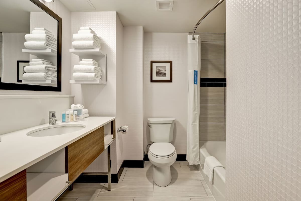 A modern bathroom with a white sink, toilet, and shower. There are towels stacked on shelves and a framed picture on the wall.