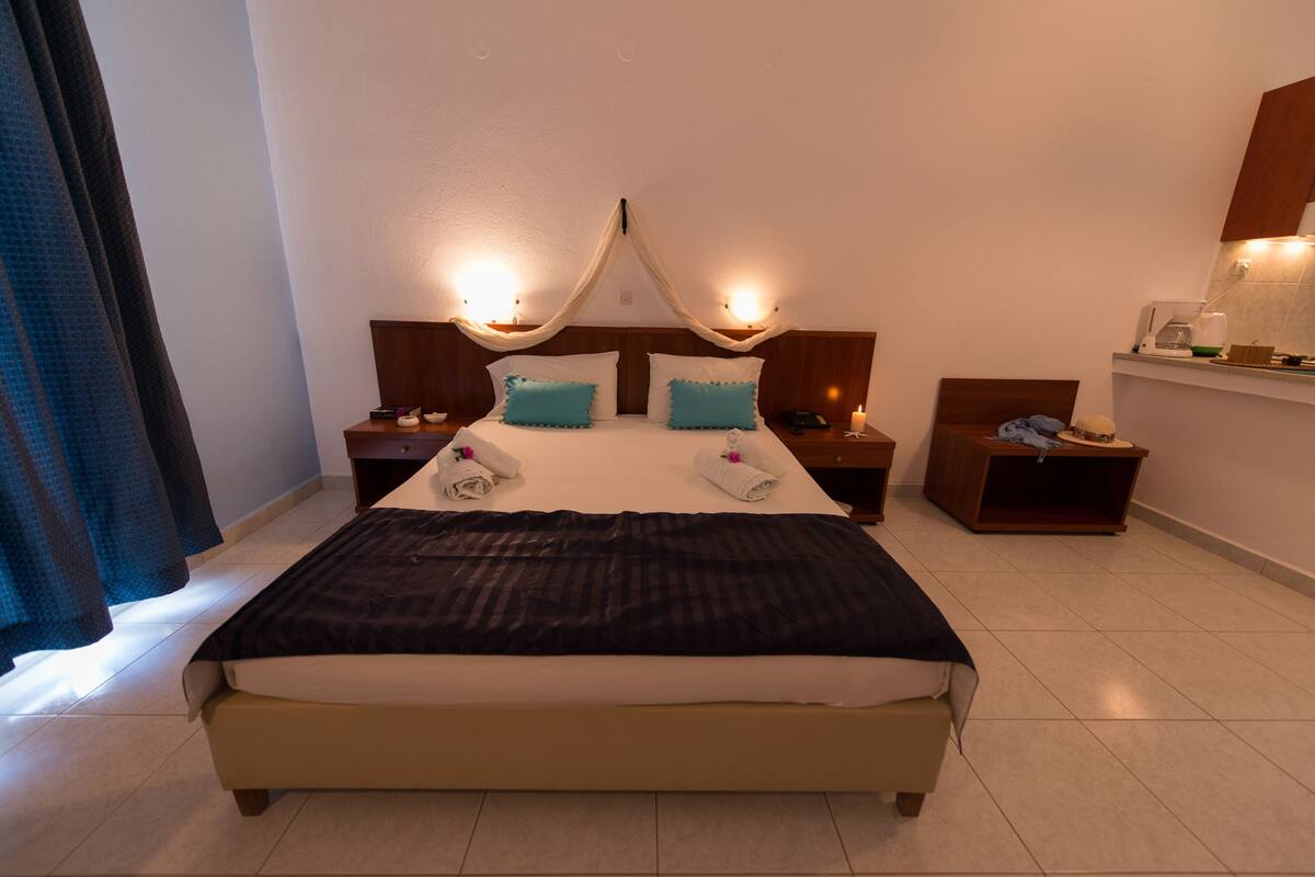 Una cama bien hecha con un edredón oscuro, dos almohadas y un dosel sobre ella. Hay mesitas de noche con lámparas y un pequeño estante con objetos.