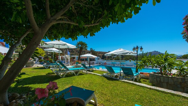 Un área de piscina con sillones de descanso y sombrillas, rodeada de vegetación y un cielo despejado.