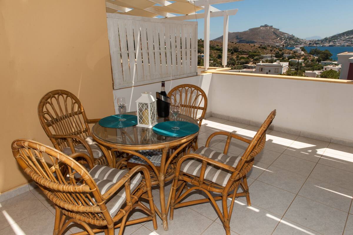Una mesa redonda de vidrio con sillas de mimbre, una botella y un vaso en un balcón con vistas a un pueblo y montañas.