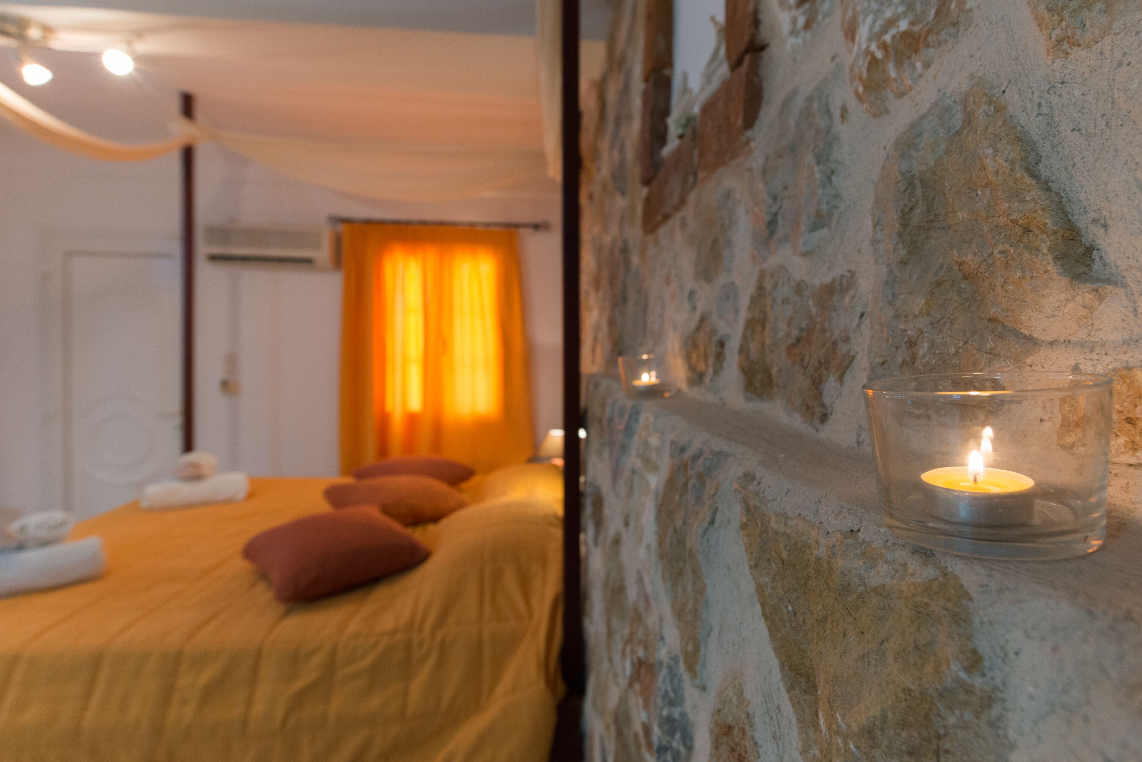 A bedroom with a stone wall, a bed with yellow bedding, and a lit candle in a glass holder.