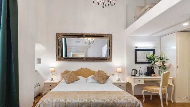A hotel room with a large bed, a desk with a chair, a mirror, and a chandelier.