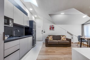 A modern kitchen with a microwave, stove, and sink. There's a brown sofa with colorful cushions in a living area with a wooden floor.