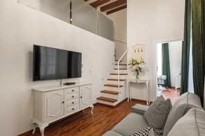 Een moderne woonkamer met een flatscreen televisie, een witte console, een trap en een bank met opvallende kussens.