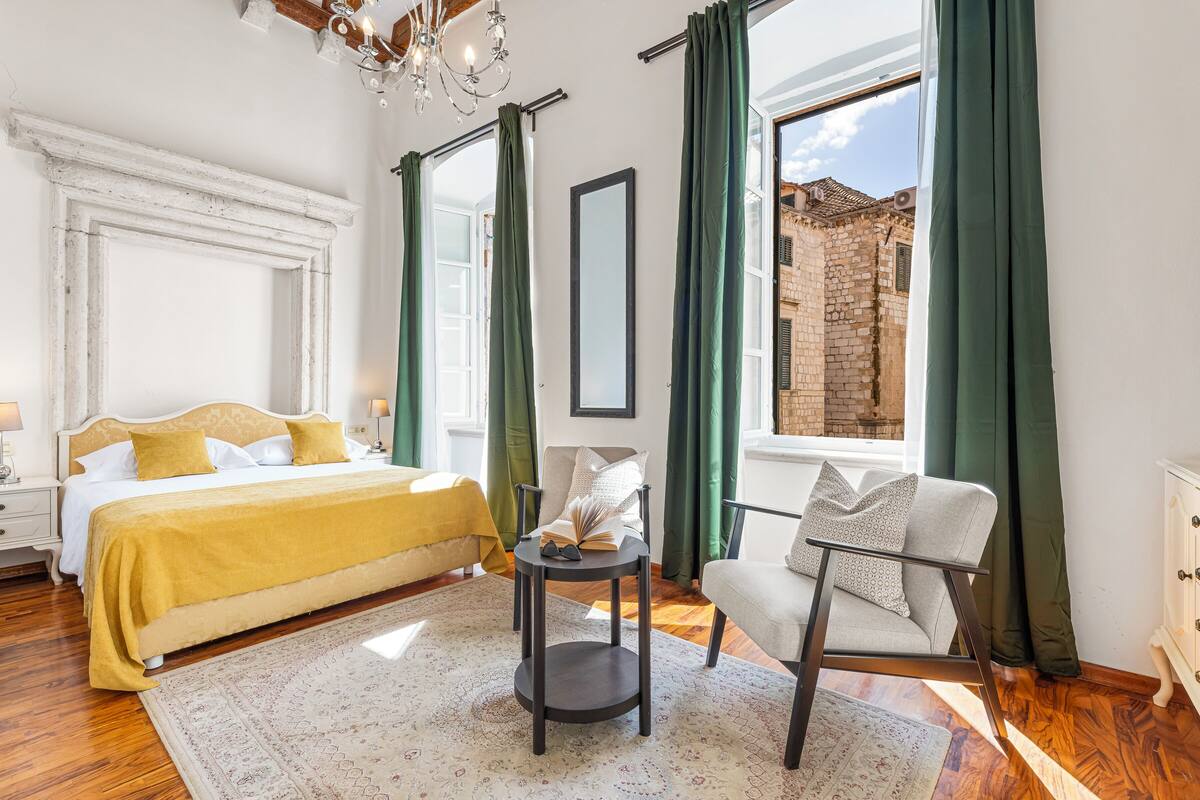 Une chambre à coucher comprenant un lit, un fauteuil de lecture, une petite table et une vue sur un bâtiment historique par la fenêtre.