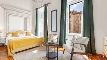 A bedroom with a bed, a reading chair, a small table, and a view of a historic building through the window.