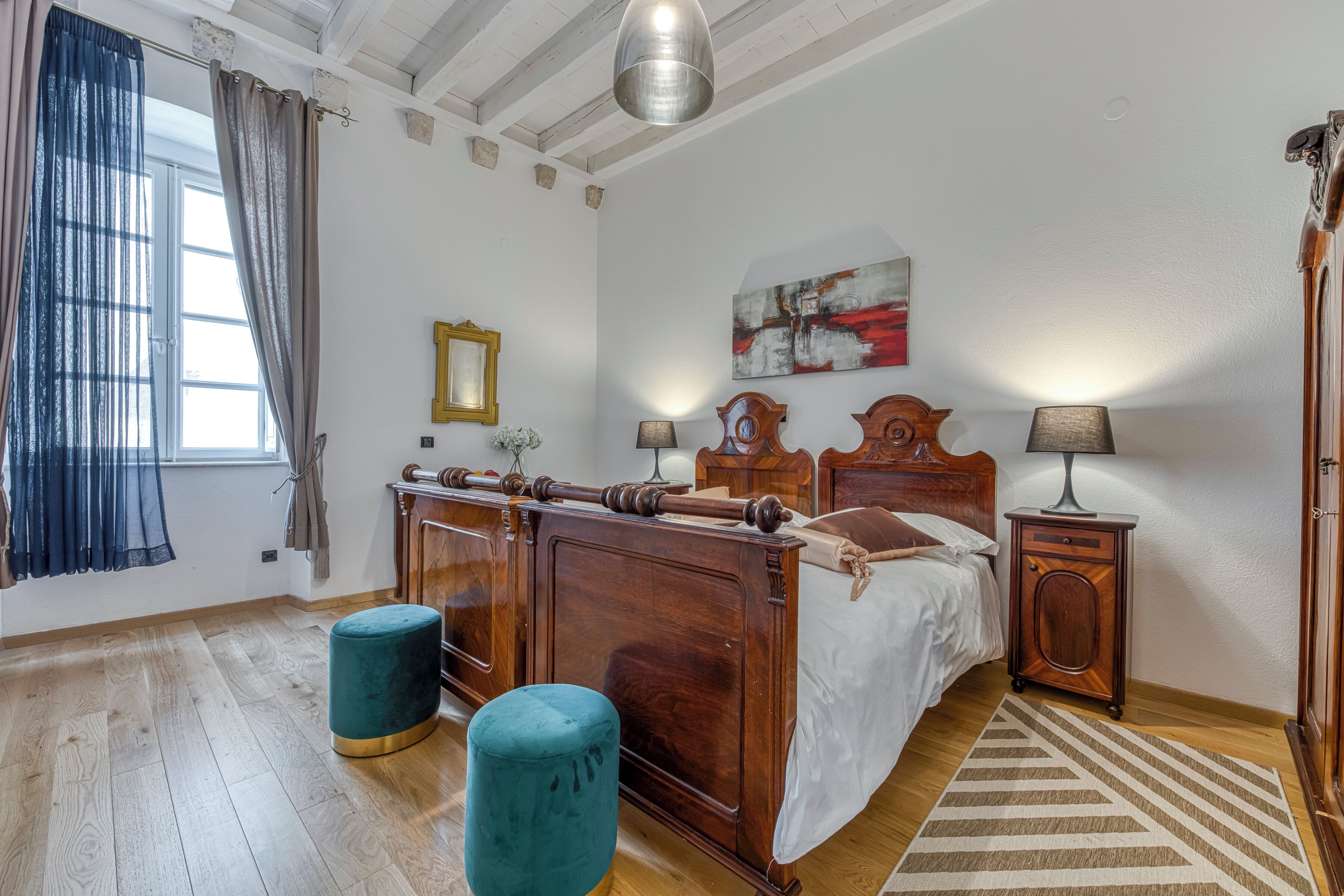A bedroom with a wooden bed, a nightstand, a lamp, a window with curtains, and a painting on the wall.