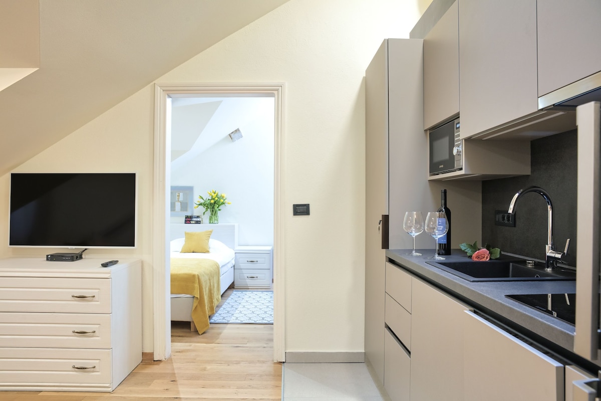 A modern kitchen with a microwave, sink, and wine glasses. There is a bed with a yellow blanket and a plant in the adjacent room.