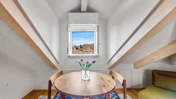 A room with a round wooden table, chairs, a window with a view of buildings, and a small vase with flowers.