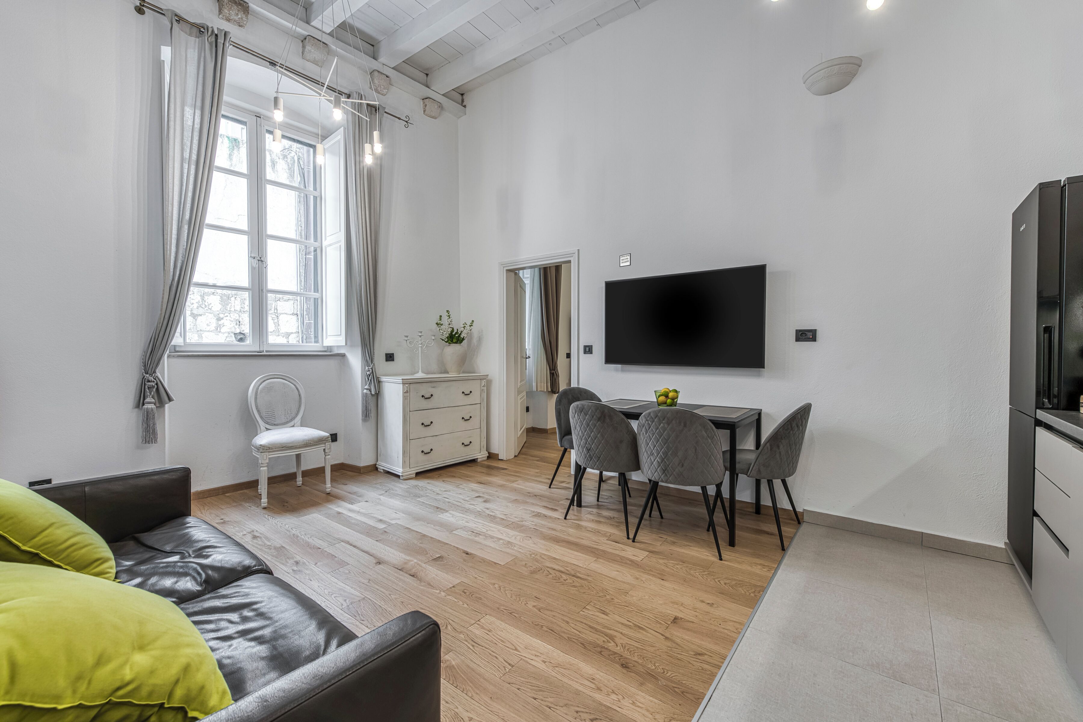 Una sala de estar moderna con un sofá, una mesa de comedor con sillas, un televisor montado en la pared y un ventanal con cortinas.