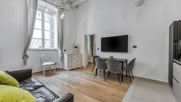 Una sala de estar moderna con un sofá, una mesa de comedor con sillas, un televisor montado en la pared y un ventanal con cortinas.