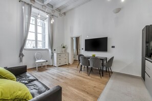 Een moderne woonkamer met een bank, een eettafel met stoelen, een aan de muur bevestigde televisie en een raam met gordijnen.