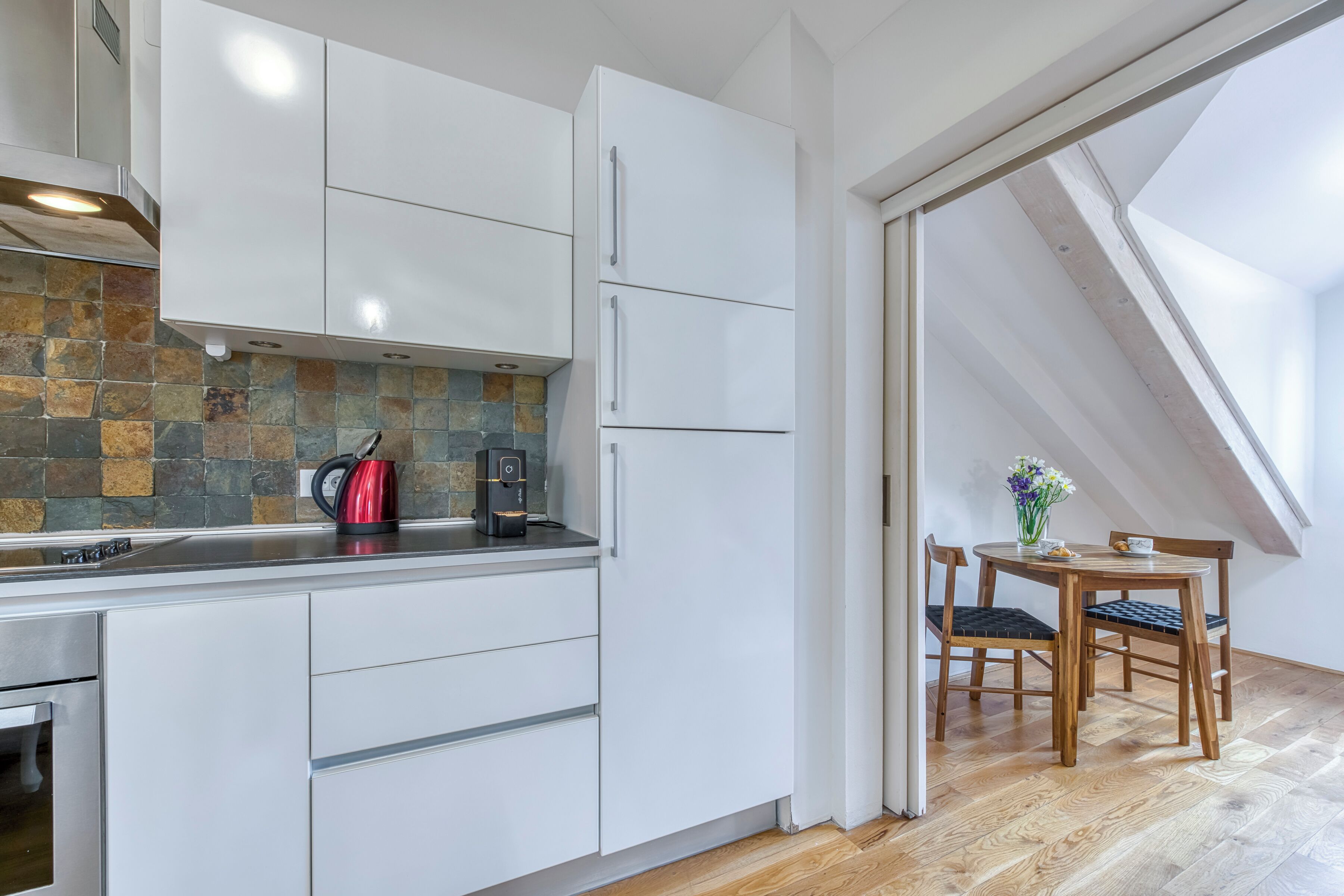 A modern kitchen with white cabinets, a stone backsplash, and a dining area with a wooden table and chairs.