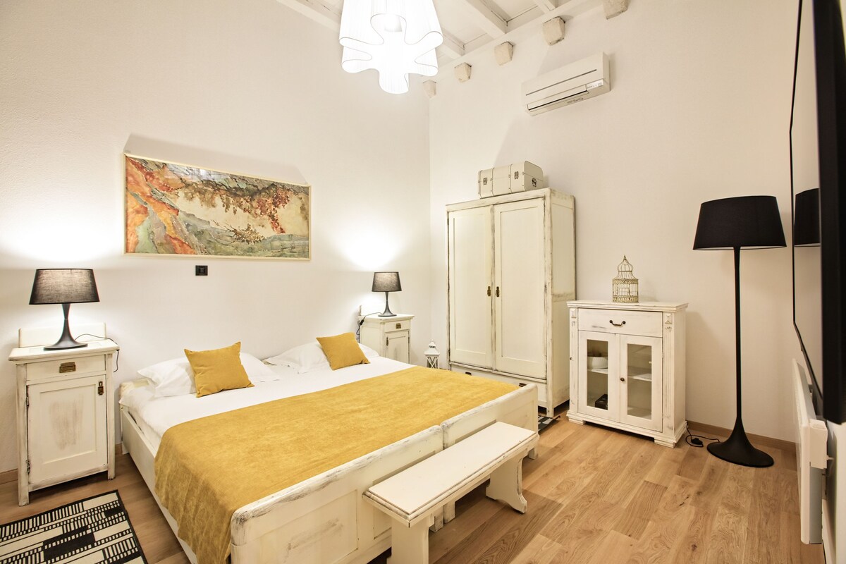 A bedroom with a bed, bedside tables, a cabinet, a bench, a floor lamp, and a painting on the wall.